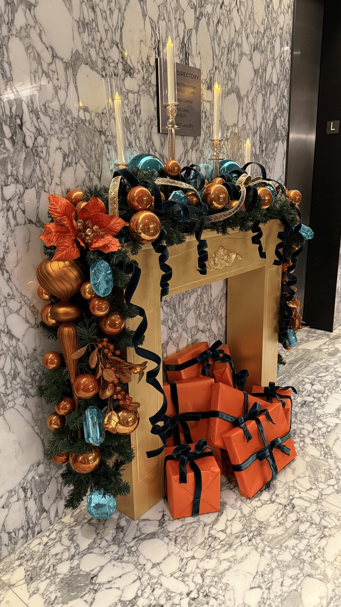 Gold mantel richly styled with copper and teal ornaments, poinsettias, candlelight, and gifts in a marble lobby