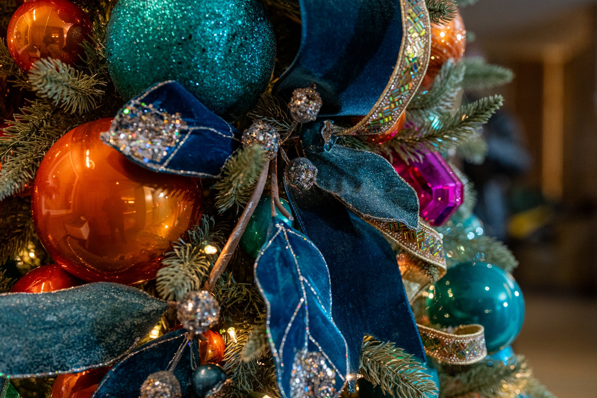Close-up of a designer Christmas tree vignette: teal velvet bows, glitter-tipped magnolia leaves, copper ornaments, and jeweled ribbon