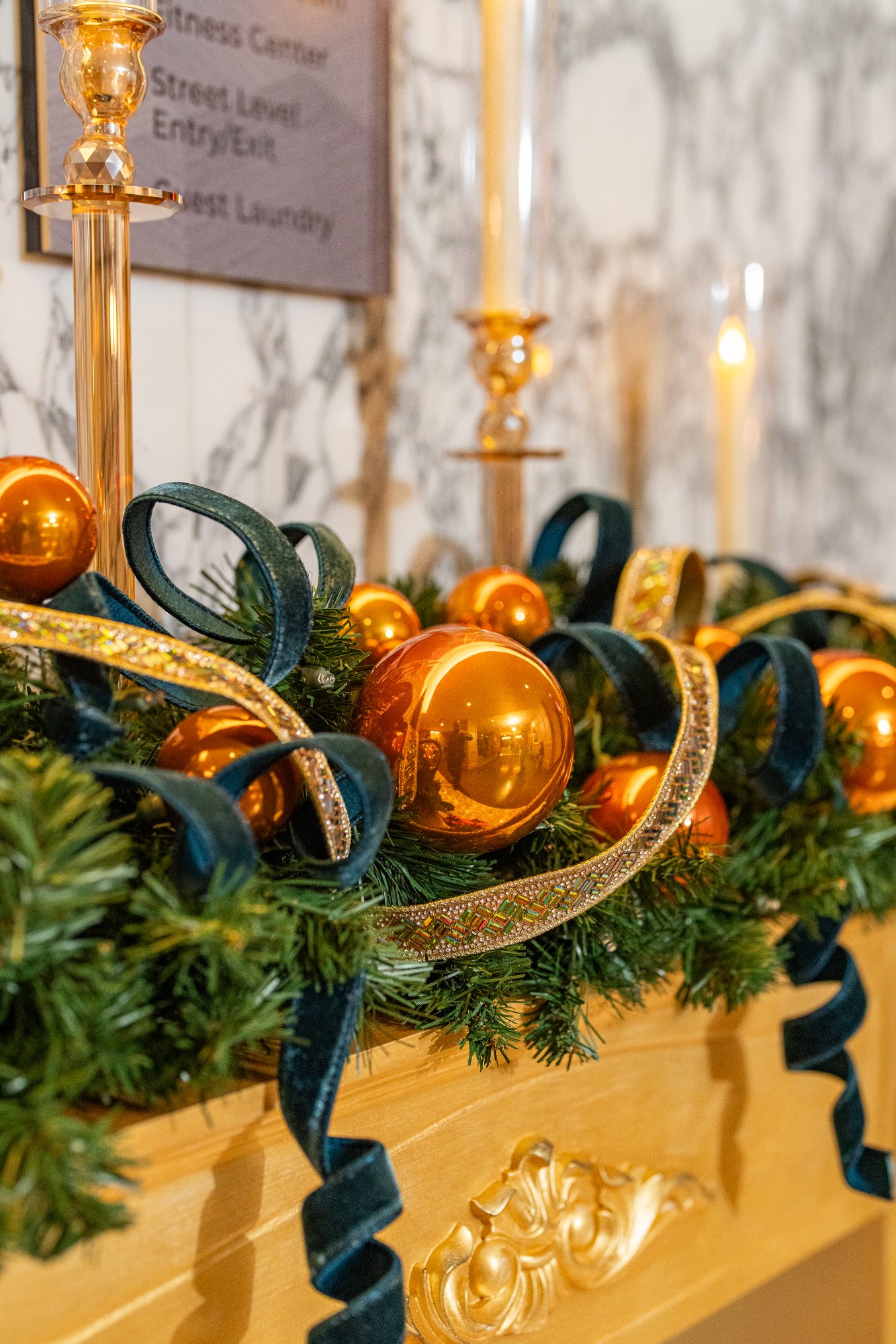 Commercial mantel garland detail: polished copper ornaments, teal velvet ribbon loops, and jeweled gold trim beside candlelight