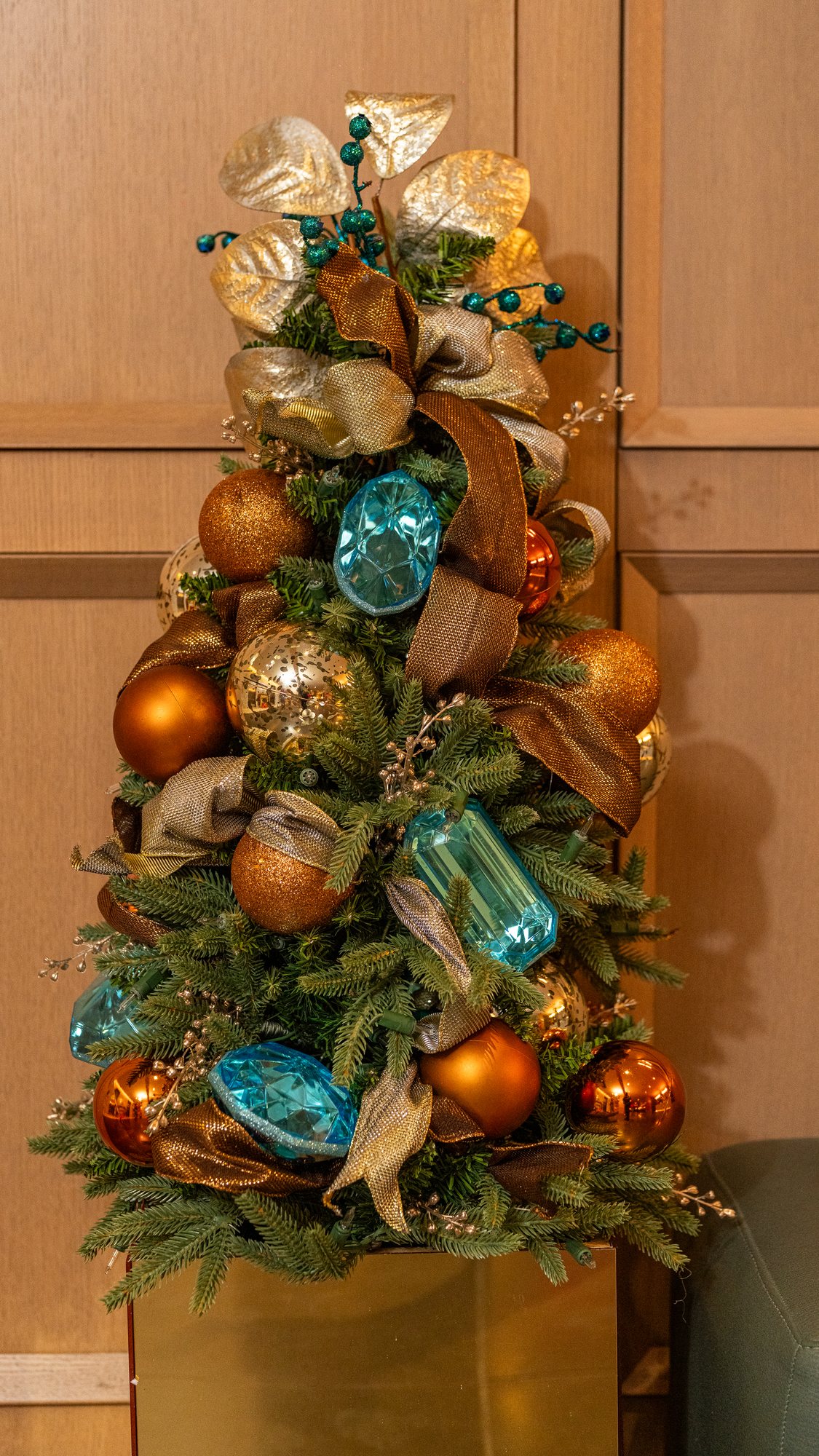 Tabletop Christmas tree in copper, gold, and aqua gems on a gold pedestal against a wood-panel wall