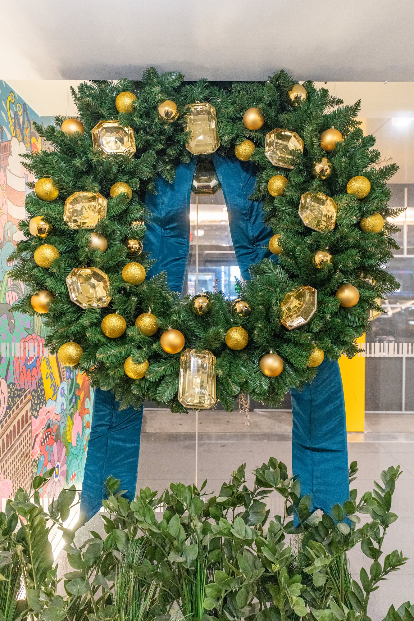 Large evergreen wreath set with amber gem ornaments and gold balls, finished with deep teal velvet ribbon tails