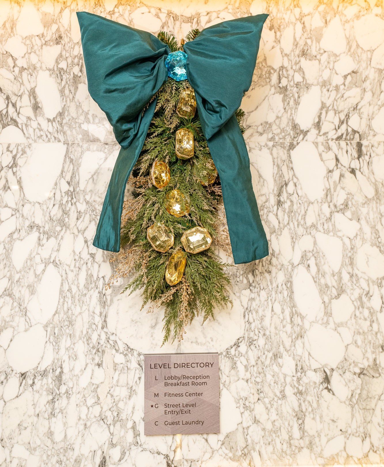 Teal velvet bow atop a cascading evergreen drop set with amber gem ornaments on a marble wall