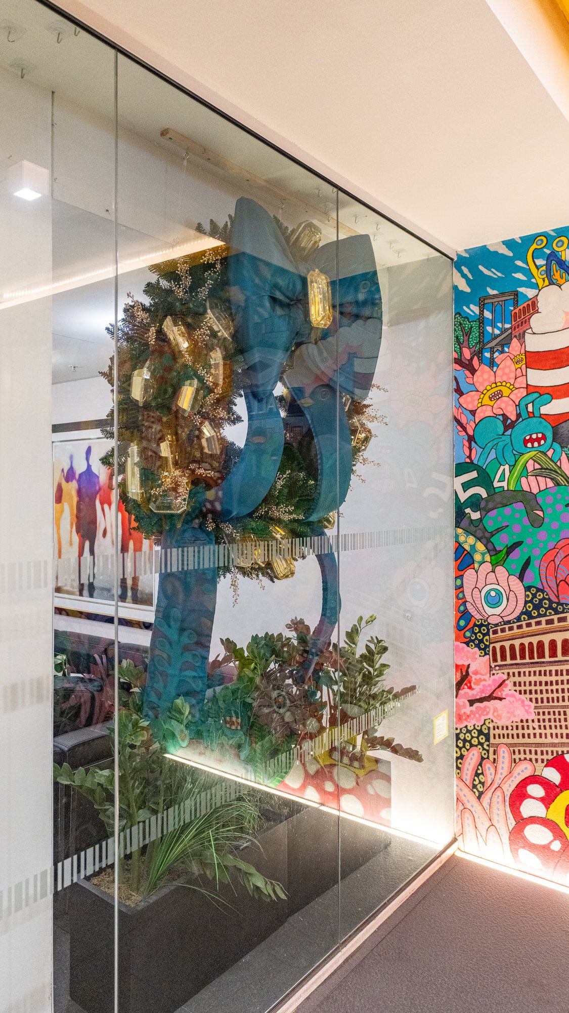 Evergreen wreath with a teal velvet bow displayed behind glass beside a colorful mural in a hotel corridor