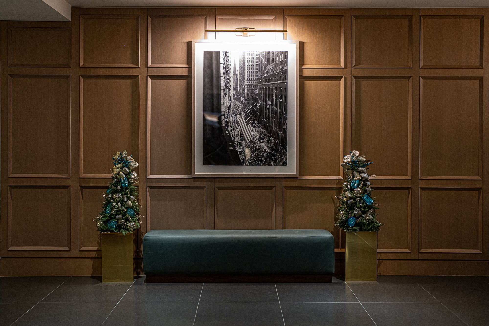 Pair of matching mini Christmas trees in teal, gold, and silver flanking a bench beneath a framed New York photograph
