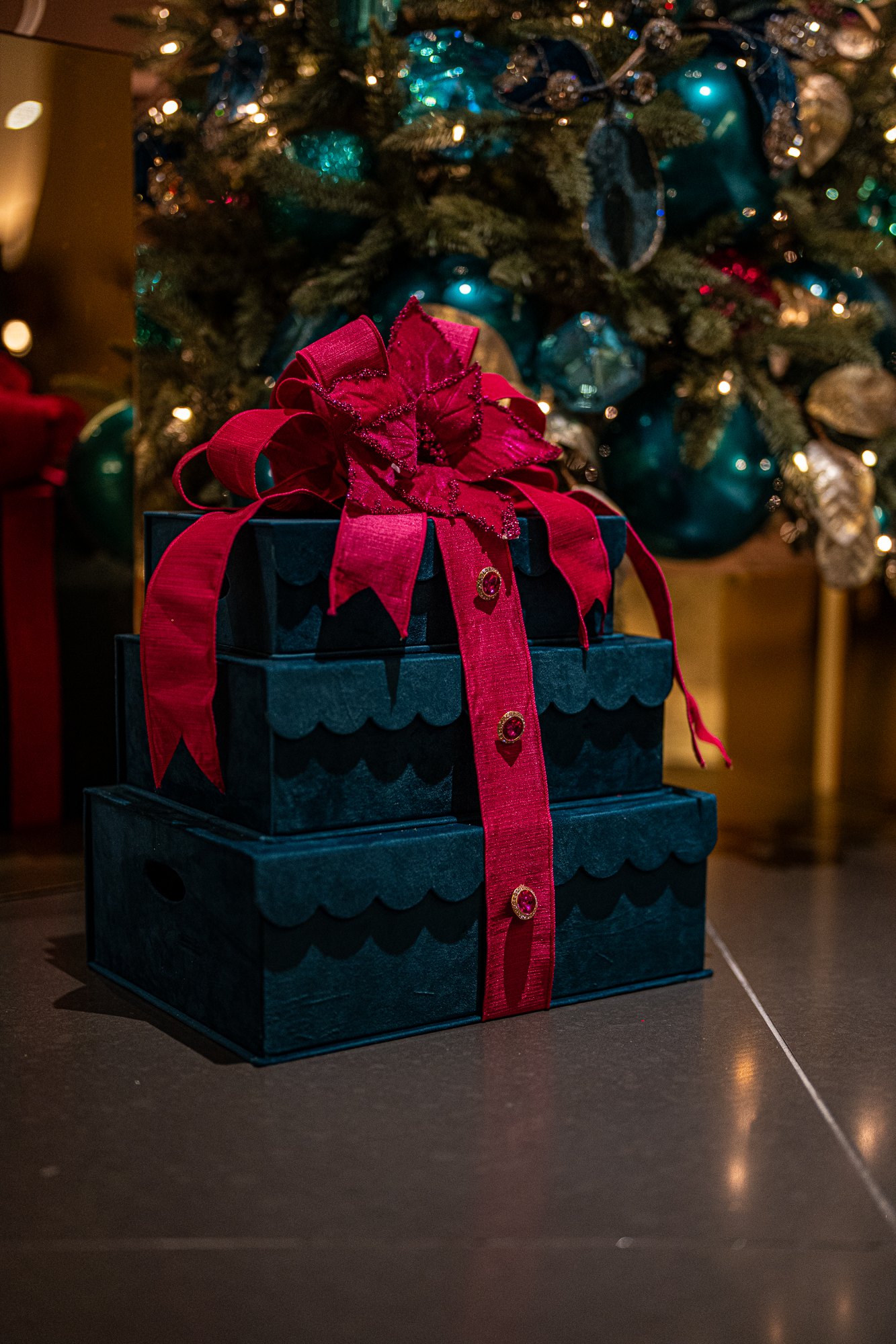 Stacked navy velvet gift boxes tied with a crimson bow beneath the jewel-tone Christmas tree