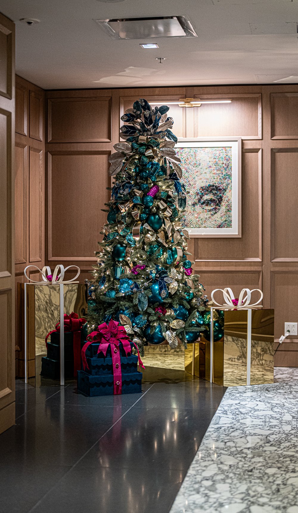 Custom jewel-tone Christmas tree in teal and gold with gem ornaments and mirrored gift boxes in an elegant paneled space