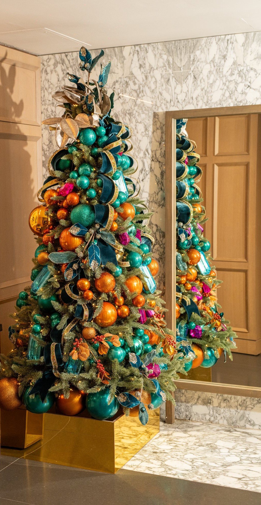 Statement corporate holiday tree in teal and copper reflected in a full-height mirror within a marble lobby