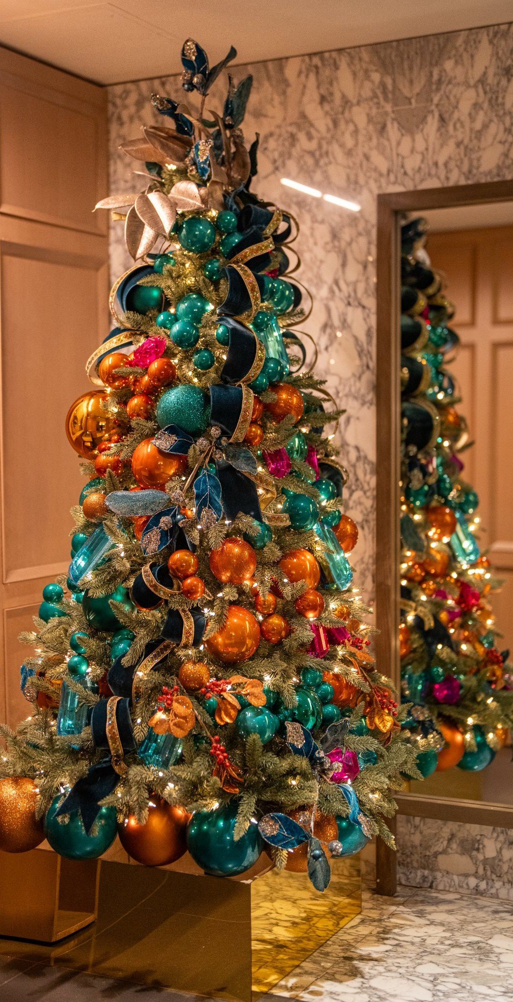 Floor-to-ceiling luxury Christmas tree dressed in teal, copper, and jewel-tone ornaments with velvet ribbon, installed in an elegant marble lobby